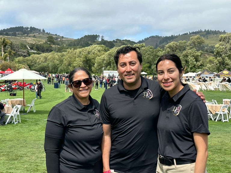 Three people in black shirts smiling at an outdoor event with green hills, white tents, and crowds in the background